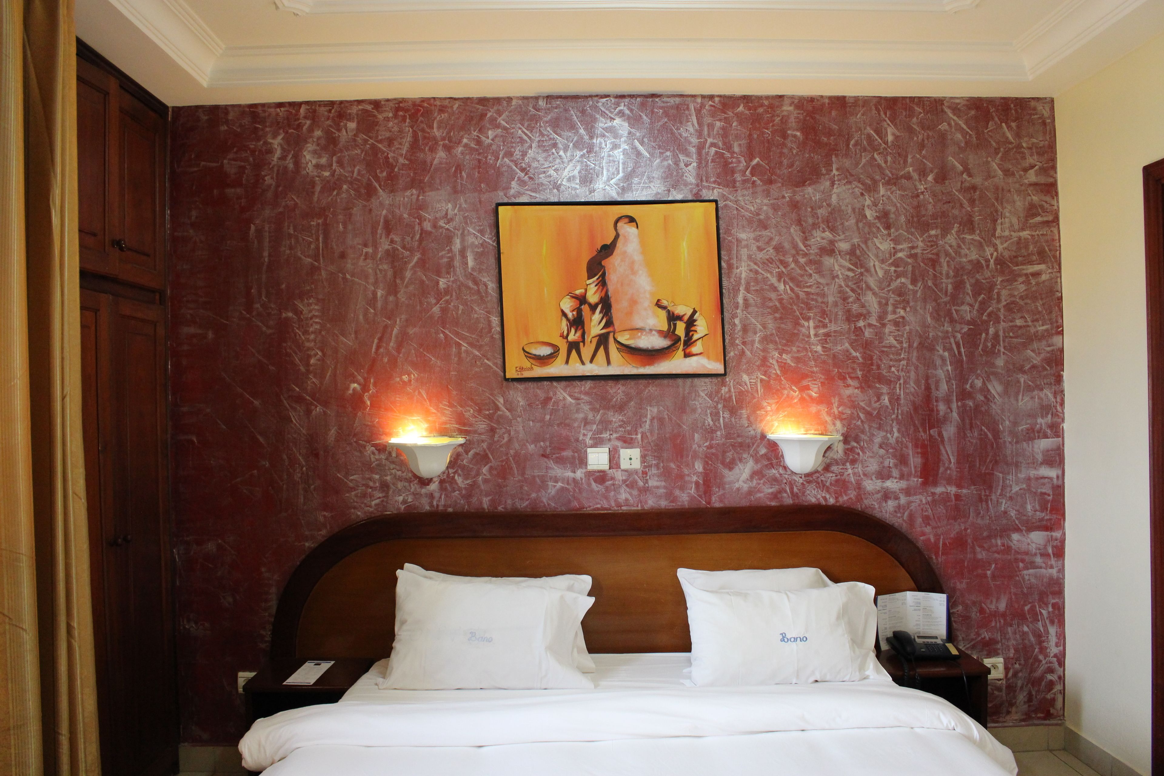 Habitación de hotel con pared de diseño de mármol, un cuadro de una mujer y un niño, dos lámparas de noche, cabecera de madera y cama con ropa blanca.