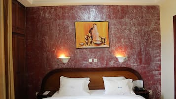 Habitación de hotel con pared de diseño de mármol, un cuadro de una mujer y un niño, dos lámparas de noche, cabecera de madera y cama con ropa blanca.