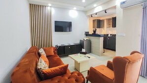 A modern living room with a sofa, armchairs, and a coffee table. A kitchen is visible in the background with a flat-screen TV mounted above the counter. - Hôtel Bano Palace (Douala)
