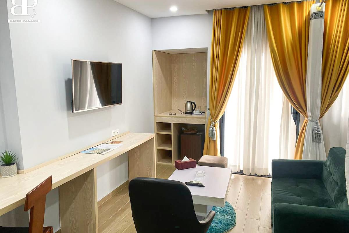 A compact hotel room with a wooden desk, a black chair, a green sofa, a TV mounted on the wall, and a small kitchenette area.