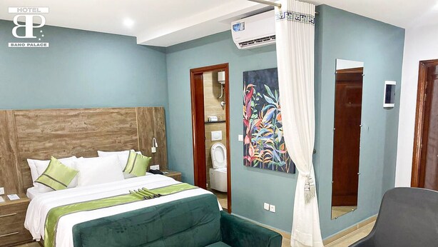 A hotel room with a bed, a green armchair, a black chair, a painting, and an air conditioning unit. - Hôtel Bano Palace (Douala)