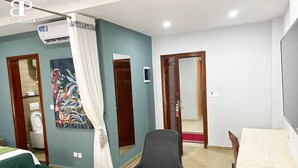 A hotel room with a bed, a desk, a chair, a painting, and a bathroom with a toilet and shower. - Hôtel Bano Palace (Douala)