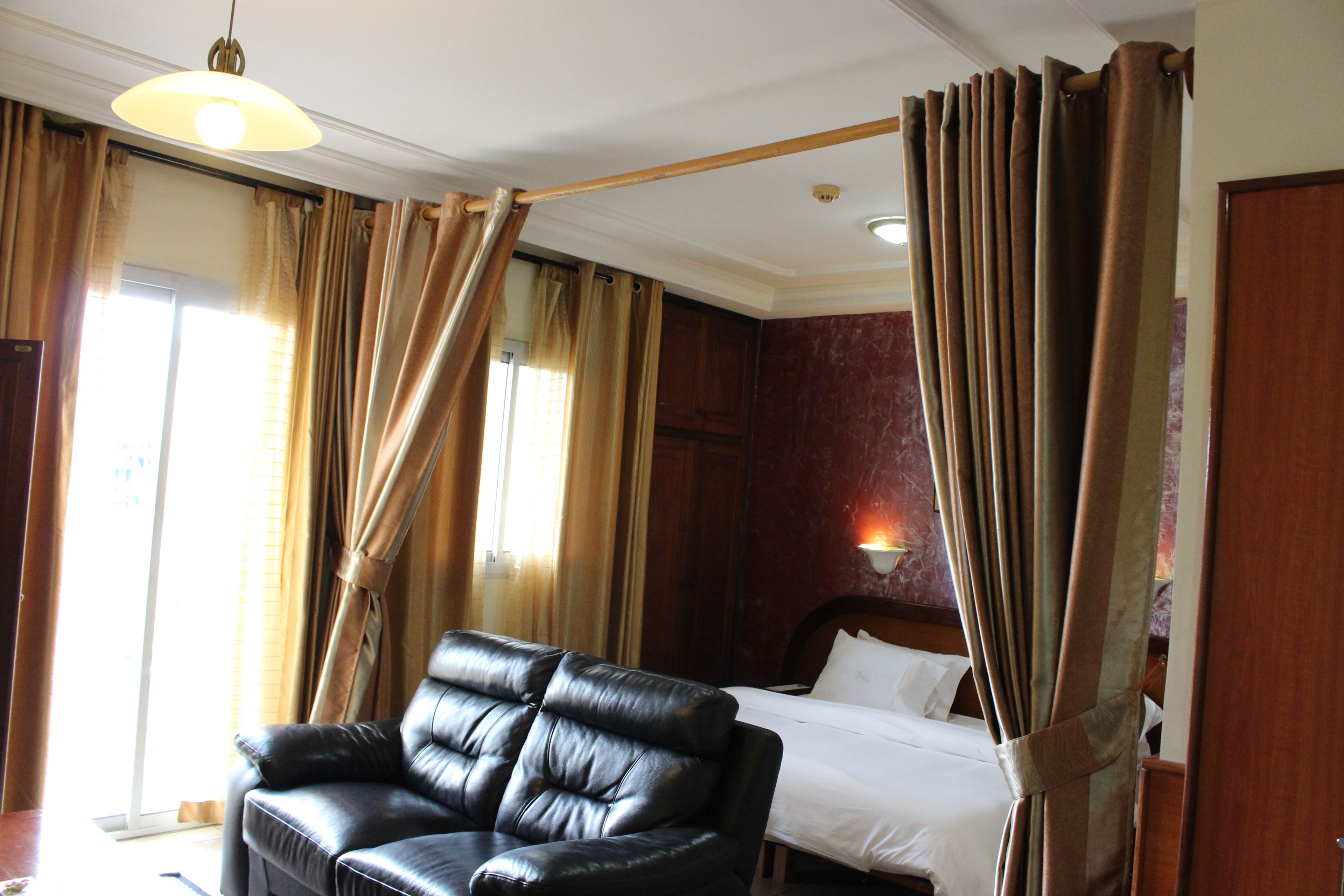 Une chambre d’hôtel avec un canapé en cuir, un lit, des rideaux et une lampe fixée au mur.