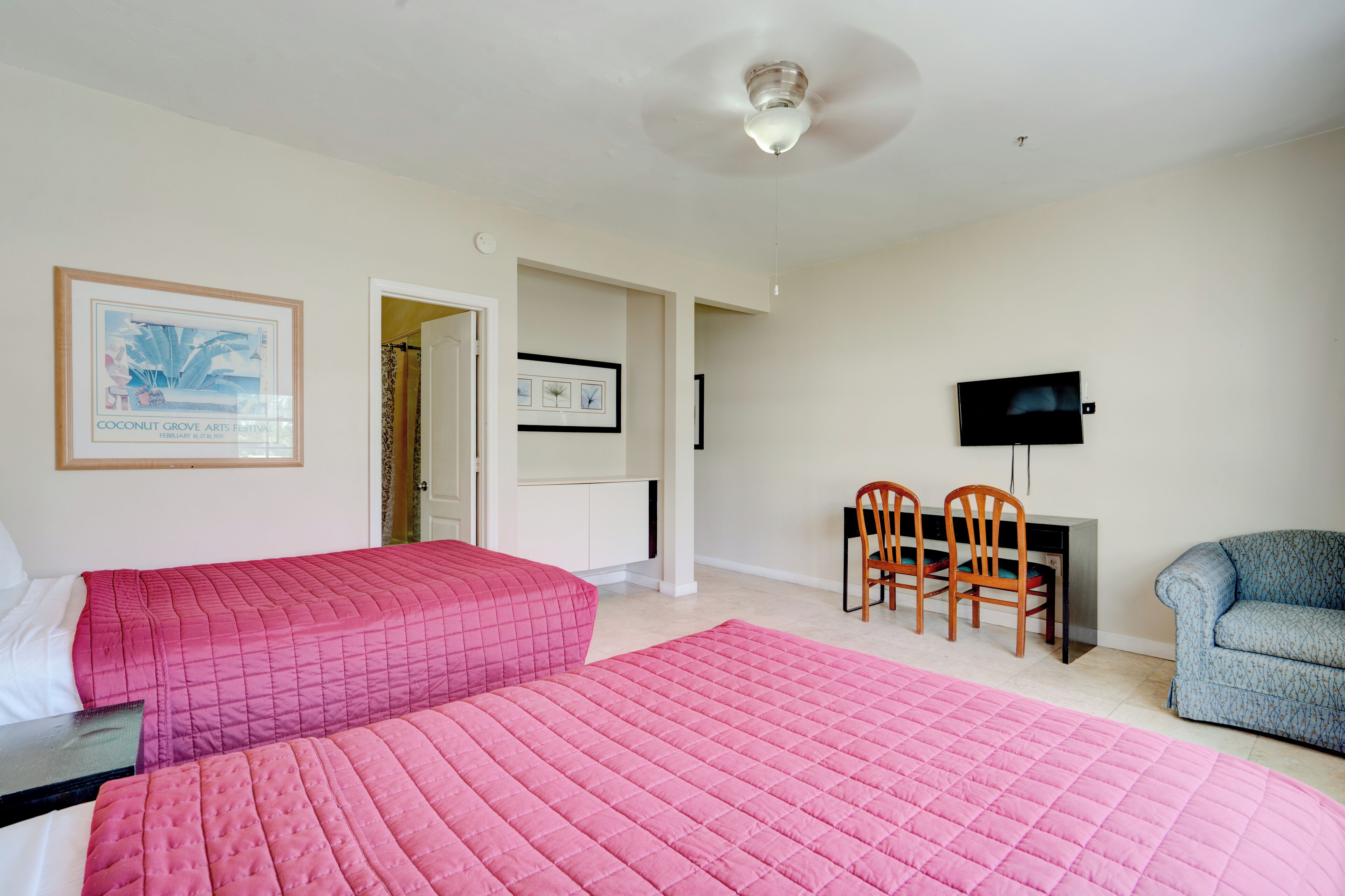 Ein Hotelzimmer mit zwei Betten, einem Schreibtisch, einem Sessel, einem Fernseher und einem gerahmten Bild an der Wand.