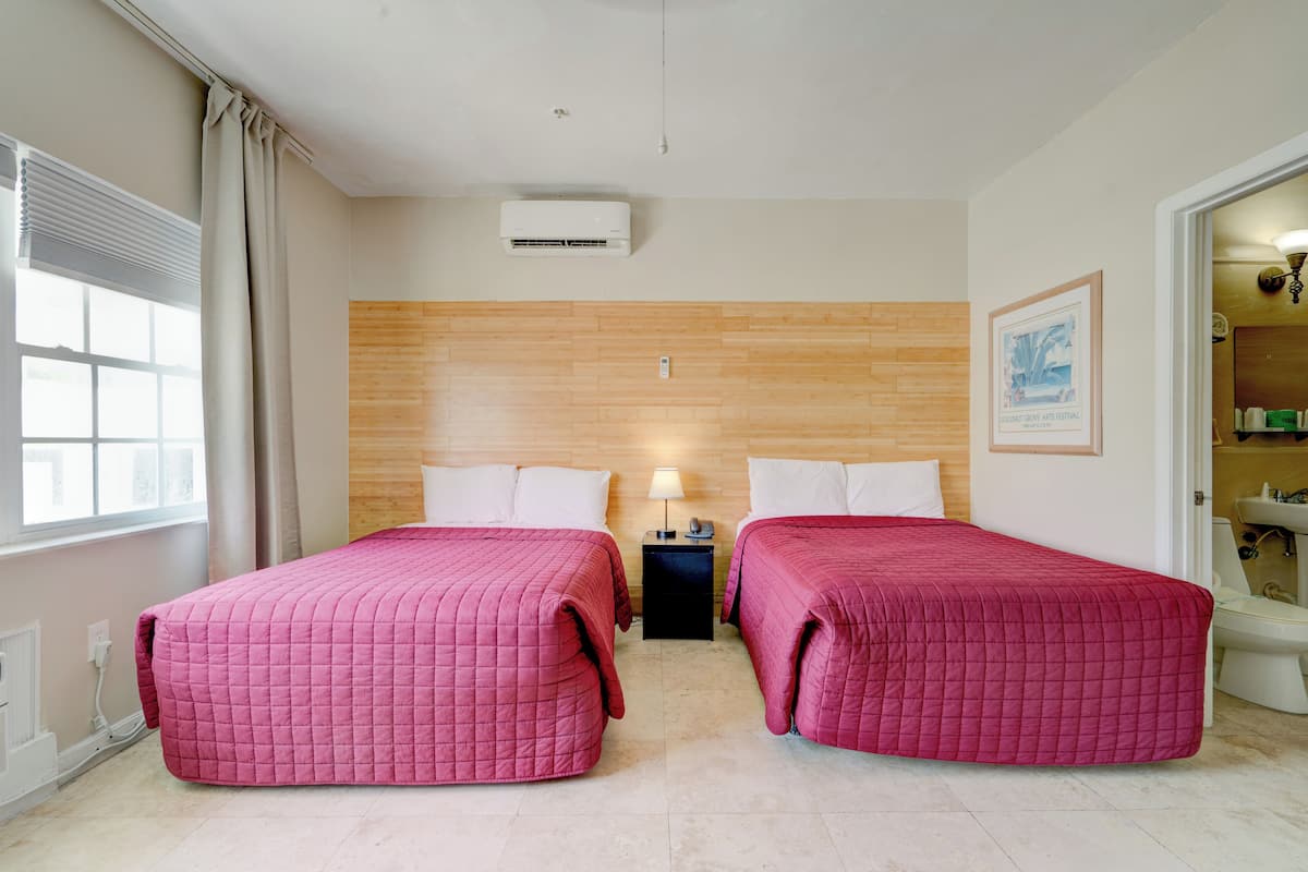 A hotel room with two beds, a wooden headboard, a window with curtains, an air conditioning unit, a nightstand with a lamp, and a framed picture on the wall.