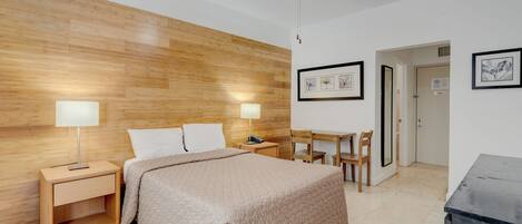 A bedroom with a wooden headboard, a bed with a beige quilt, two bedside tables with lamps, a desk with chairs, and framed pictures on the wall.