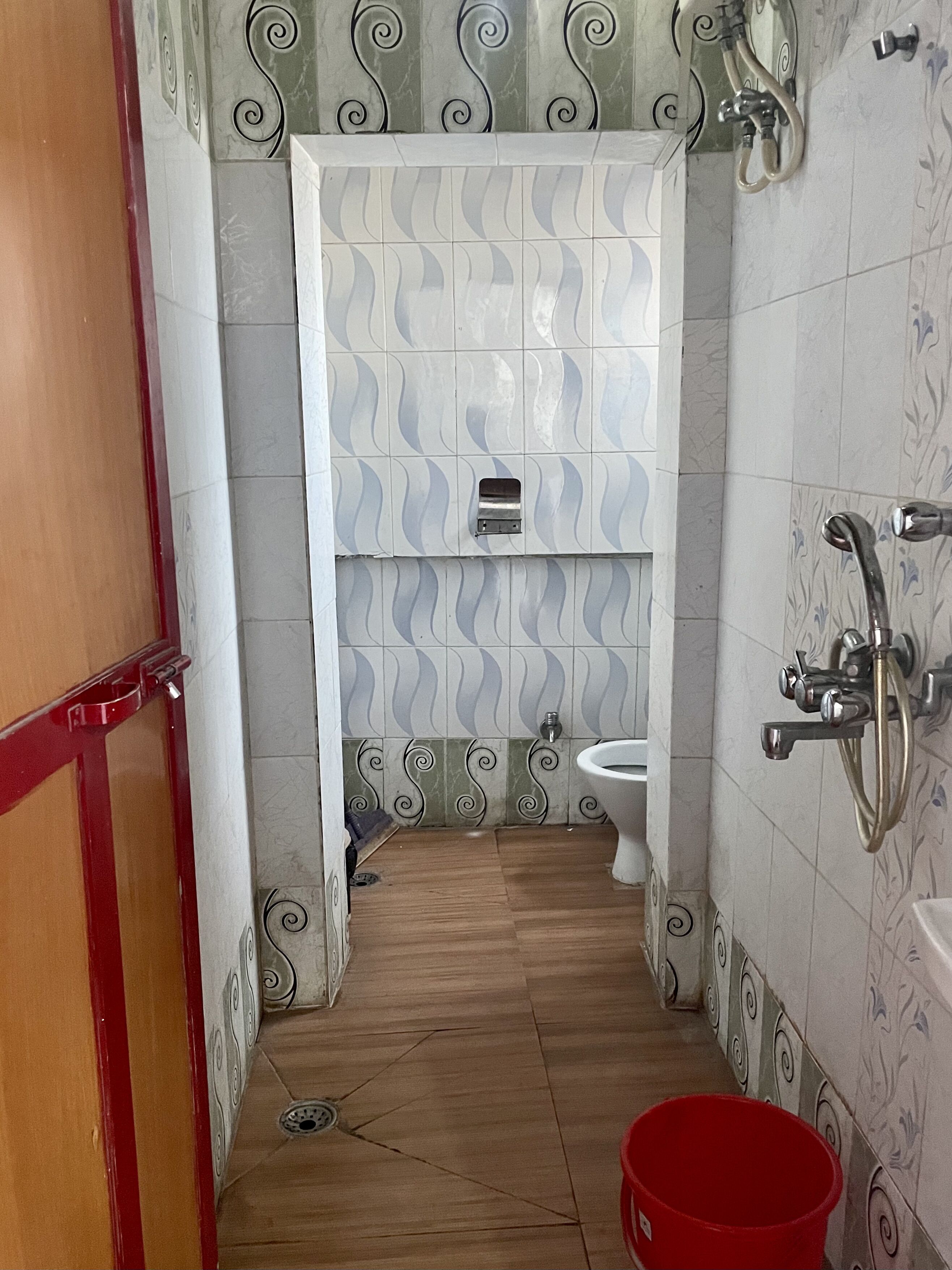 A narrow bathroom with a toilet, a red bucket, and a shower area.