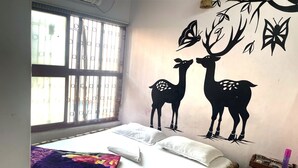 A bedroom with a bed, pillows, a blanket, a window with bars, and a wall decoration of deer and trees. - Shri Guest House (Varanasi)
