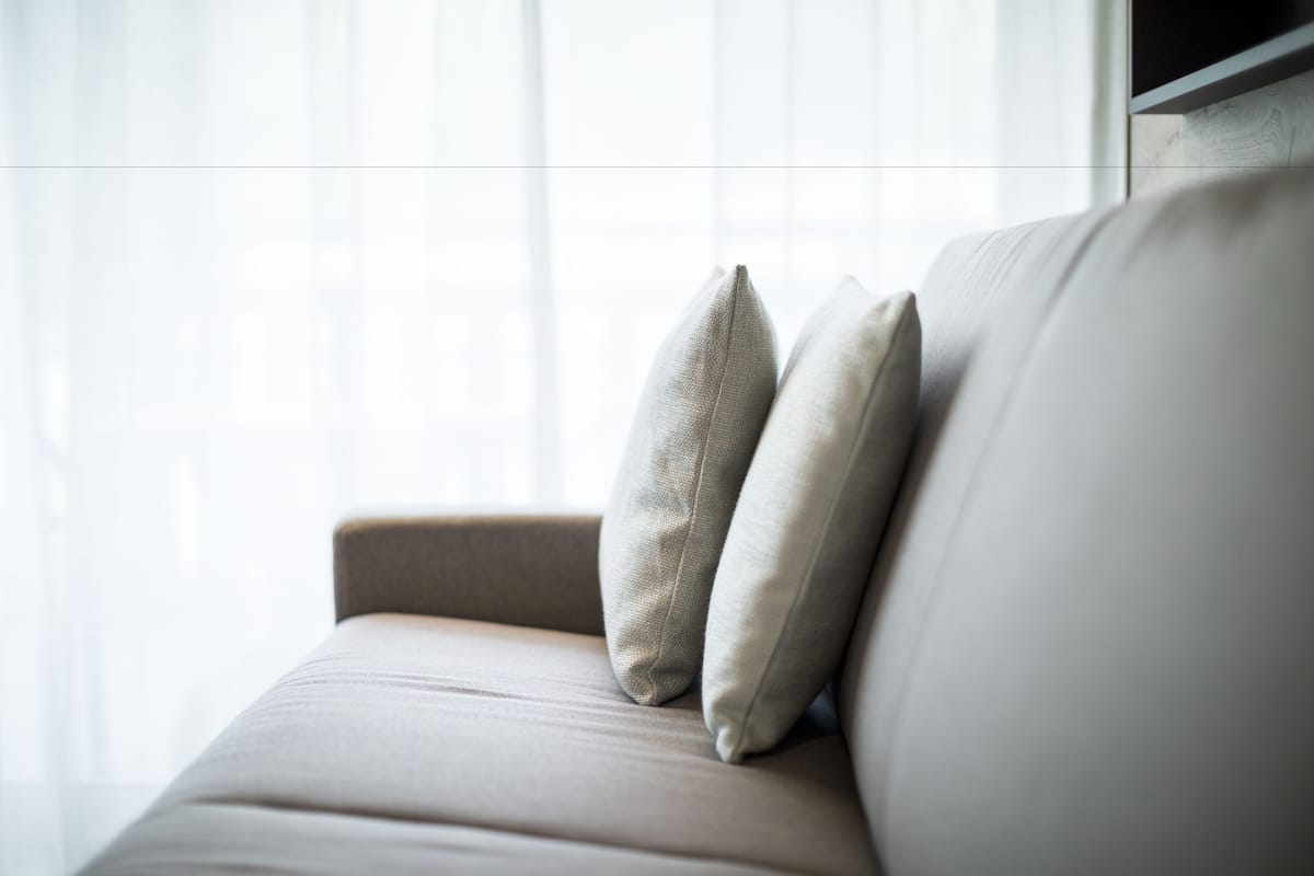 A beige sofa with two pillows against a window with sheer curtains.