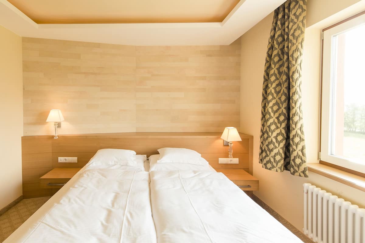 A hotel room with a wooden headboard, a double bed, two bedside tables with lamps, and a window with patterned curtains.