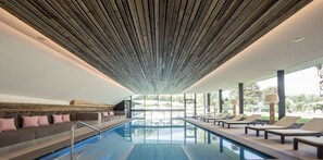 A modern indoor pool area with a wooden ceiling, lounge chairs, and a seating area with cushions.