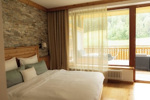 A bedroom with a large bed, a stone wall, a wooden headboard, a window with sheer curtains, and a view of a balcony.