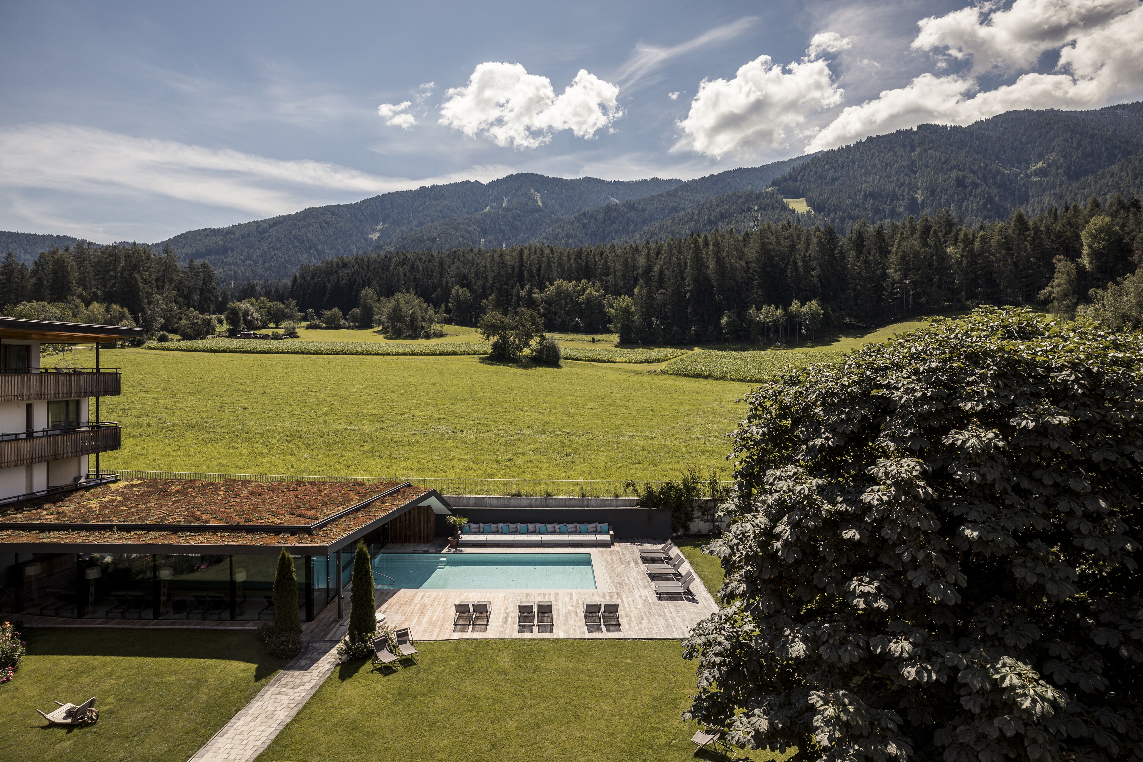 A modern building with a swimming pool and outdoor seating area, surrounded by a lush green landscape and mountains in the background.