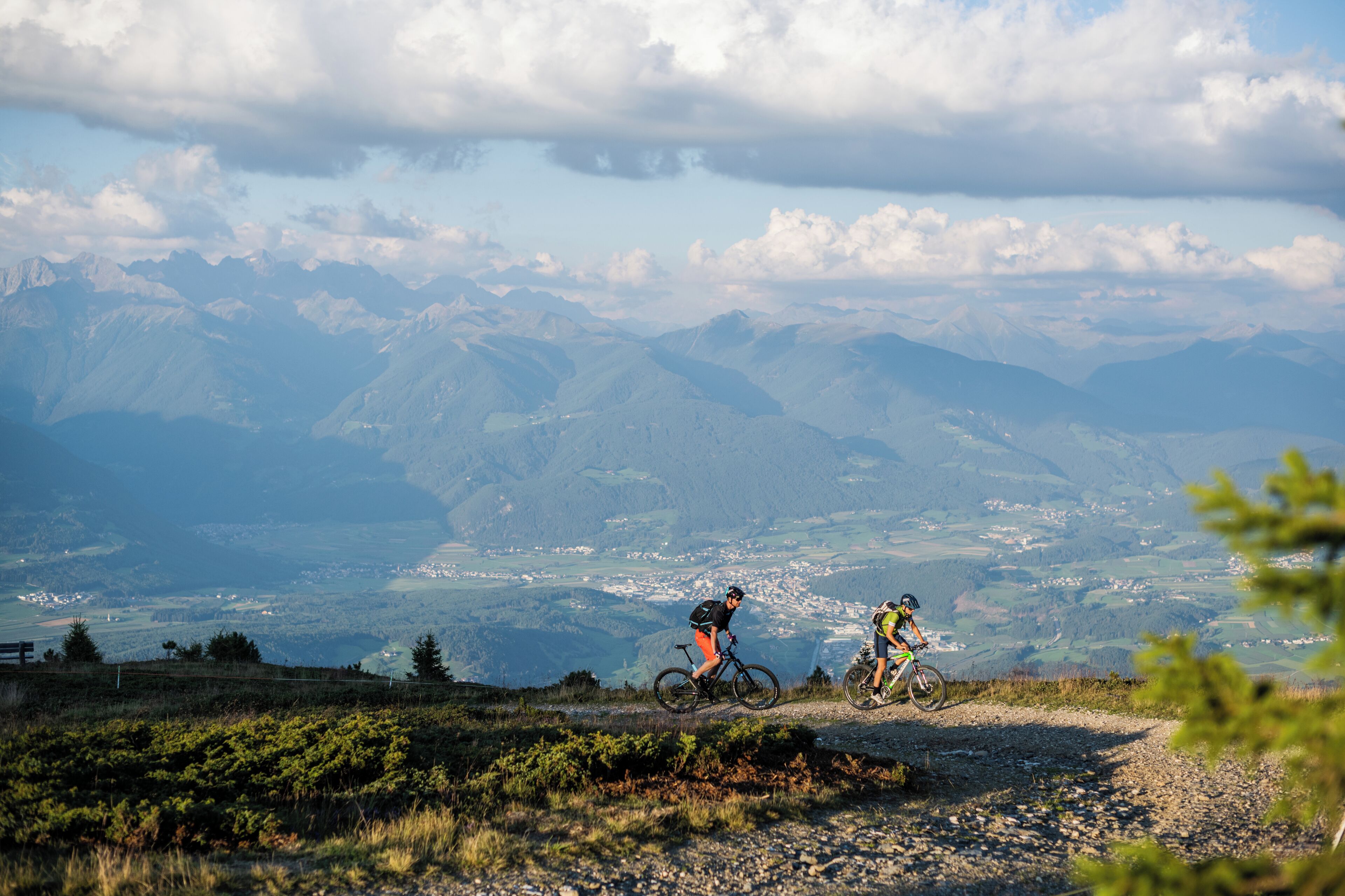 Dos ciclistas en un sendero de montaña con vistas panorámicas de montañas distantes y un valle.