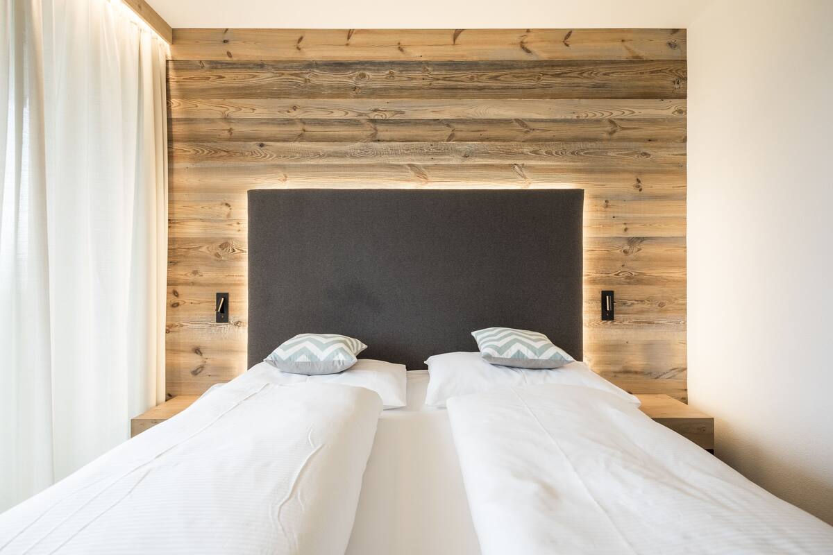 A double bed with a wooden headboard and two pillows, a bedside table, and a window with curtains.
