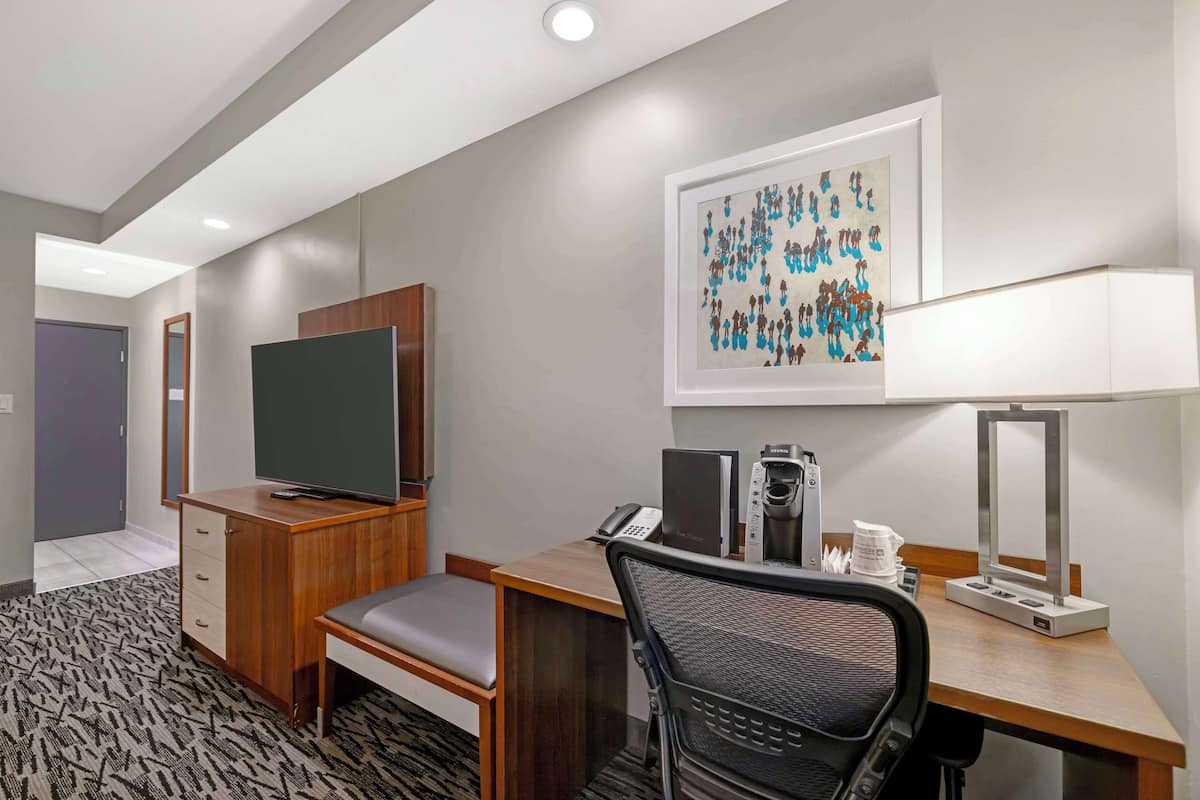 Habitación de hotel con un escritorio de madera, una silla, un televisor, un teléfono, una cafetera y un cuadro abstracto enmarcado en la pared.