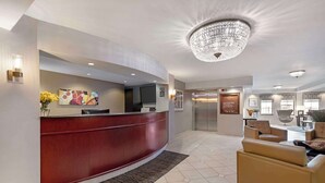 Un hall d’hôtel doté d’un comptoir d’accueil incurvé, d’un lustre, d’un espace salon avec des fauteuils en cuir et d’un sol carrelé.