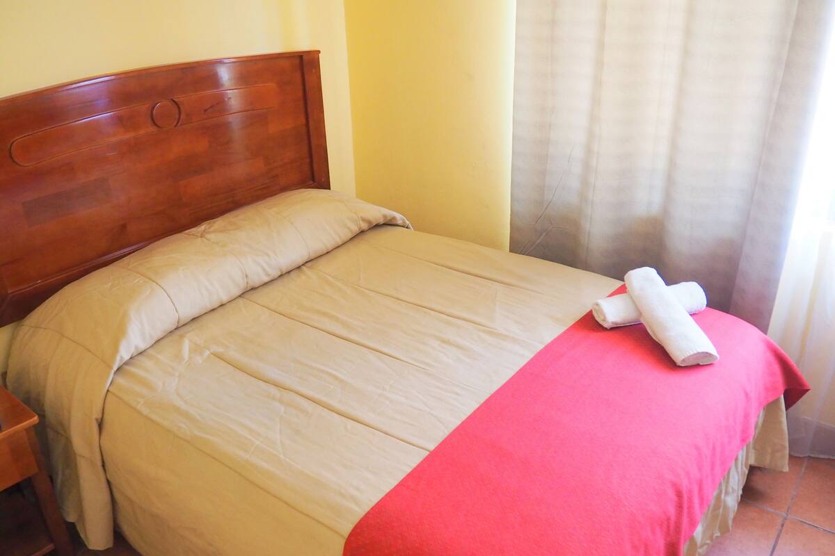 Een net opgemaakt bed met een houten hoofdbord, een beige dekbedovertrek en een rode accentkleur.