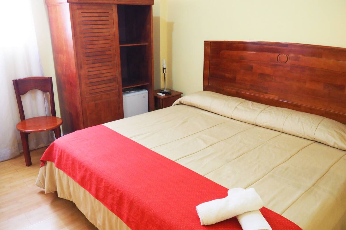 Een net opgemaakt bed met een rood-beige sprei, een houten hoofdbord en een witte handdoek er opgerold.