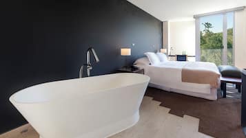 Habitación de hotel moderna con una bañera independiente grande, una cama bien hecha, una mesita de noche pequeña con lámpara y vistas a la vegetación a través de un ventanal amplio.