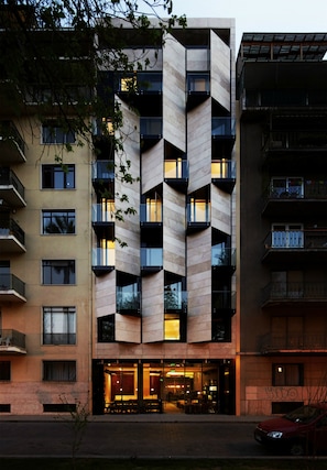 Un edificio moderno de varios pisos con una fachada geométrica distintiva, balcones y una entrada a nivel de calle con asientos.
