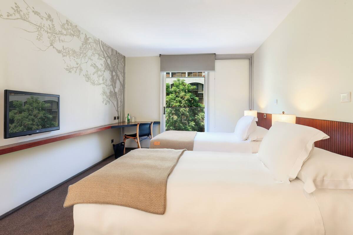 Une chambre d’hôtel avec deux lits, une télévision, un bureau et une fenêtre donnant sur des arbres.