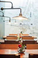 Un comedor con mesas y sillas de madera, un florero de vidrio con una sola rosa roja, y lámparas colgantes.