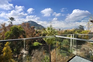A modern building with a glass balcony overlooking a park with trees and a hill in the background.