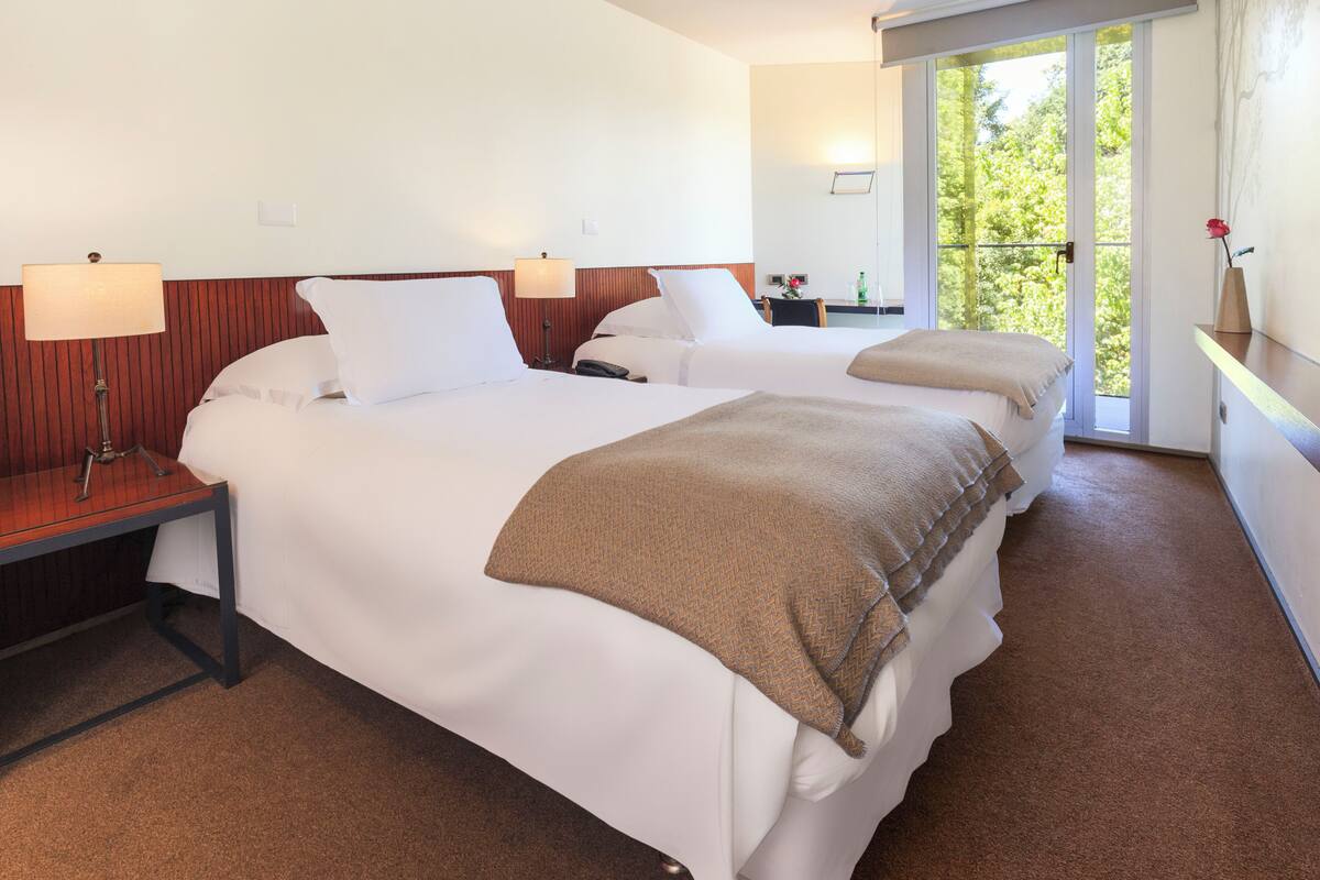Une chambre d’hôtel avec deux lits, une table de chevet, une lampe, une petite table ornée d’un vase de fleurs et une vue sur la verdure par la fenêtre.