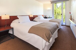 Habitación de hotel con dos camas, una mesita de noche, una lámpara, una pequeña mesa con un florero y vistas a zonas verdes por una ventana.