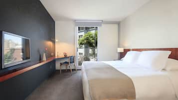 Una camera d'albergo con un letto, una scrivania con una sedia, una TV e una finestra vista edifici e alberi.