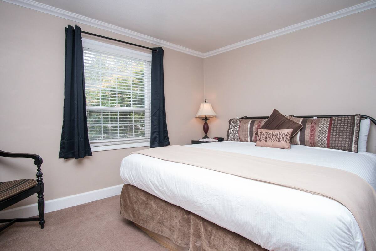 Une chambre à coucher avec un lit, une table de chevet, une lampe, une chaise et une fenêtre avec des rideaux.