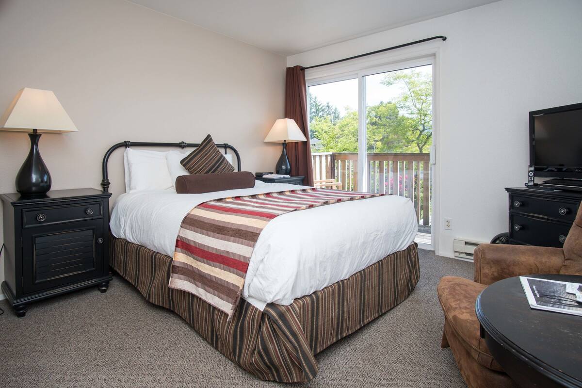 Une chambre d’hôtel comprenant un lit, deux tables de chevet avec des lampes, un téléviseur, une chaise et une fenêtre donnant sur des arbres.