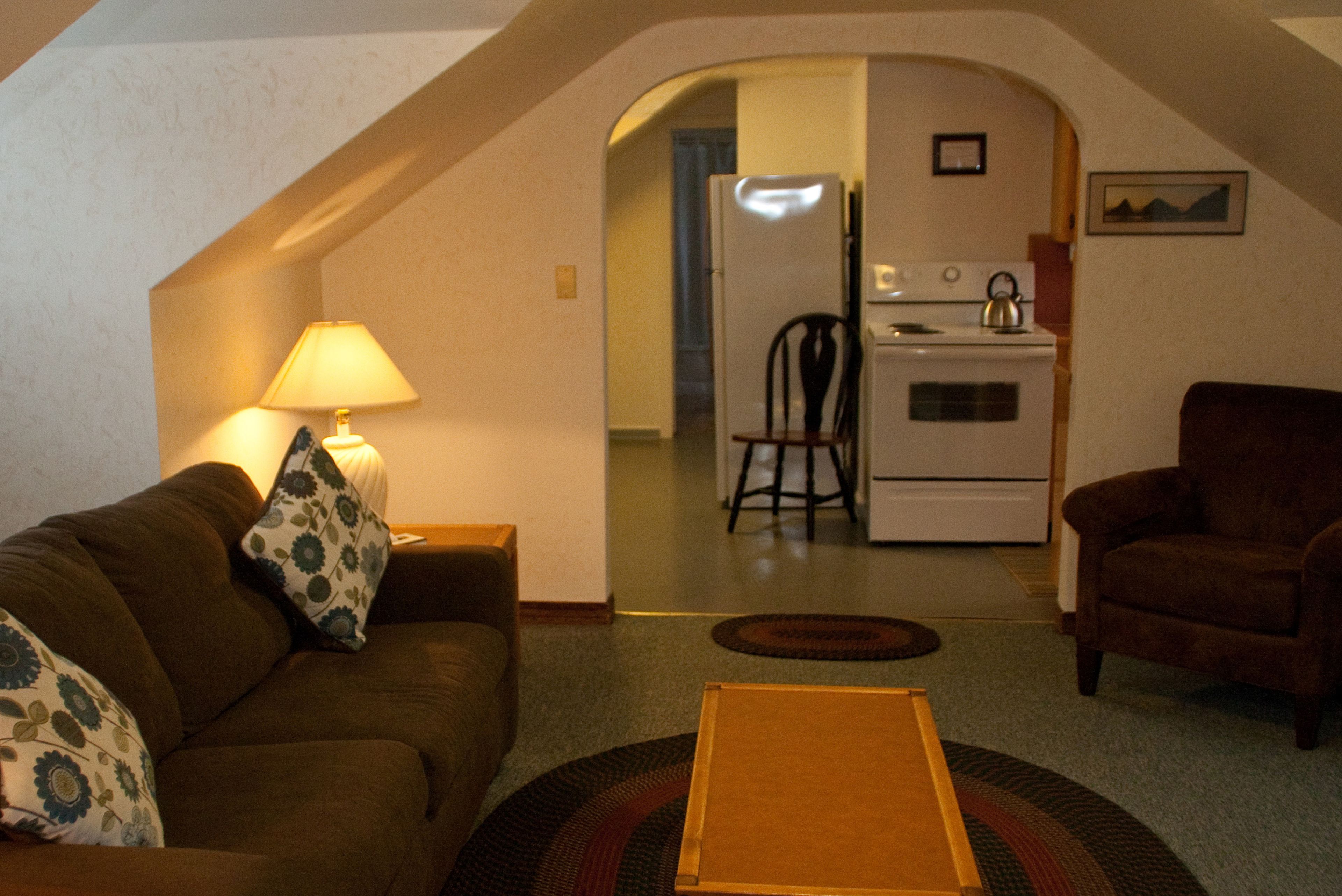 A cozy living room with a sofa, a coffee table, and a lamp. There is a kitchen area with a stove and a refrigerator in the background.