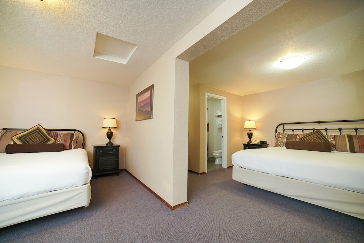 Une chambre d’hôtel avec deux lits, une table de chevet avec une lampe, un tableau au mur et une porte donnant sur une salle de bain.
