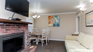 Una sala de estar con chimenea, un televisor montado encima, una mesa de comedor con sillas, un sofá color beige y un cuadro en la pared.