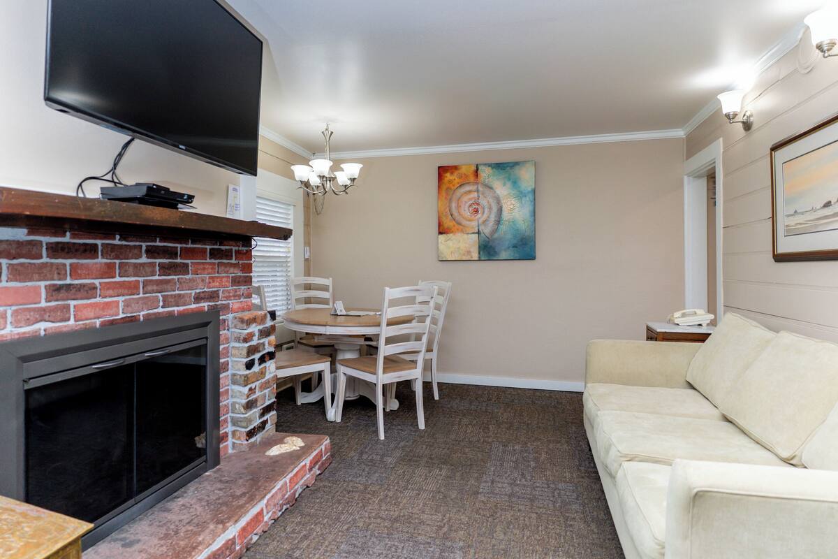 Un salon avec une cheminée, un téléviseur fixé au-dessus, une table à manger avec des chaises, un canapé beige et un tableau accroché au mur.