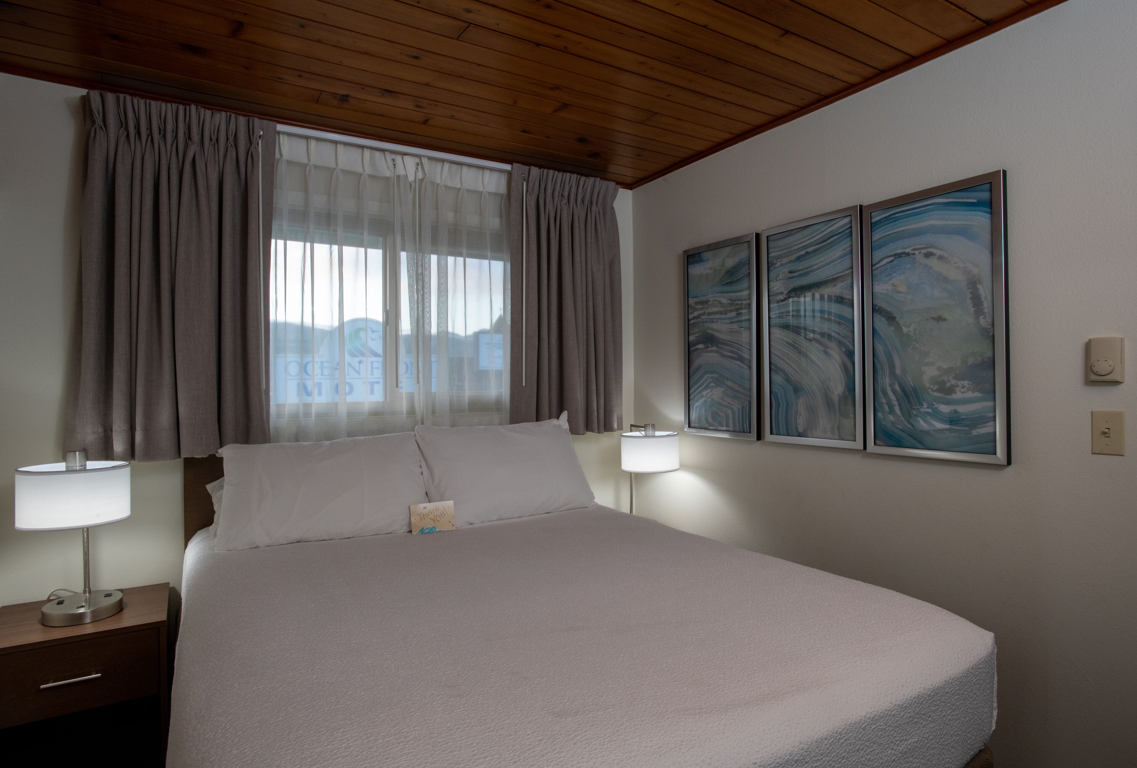 A neatly made bed in a room with wooden ceiling, two bedside lamps, and two abstract paintings on the wall.