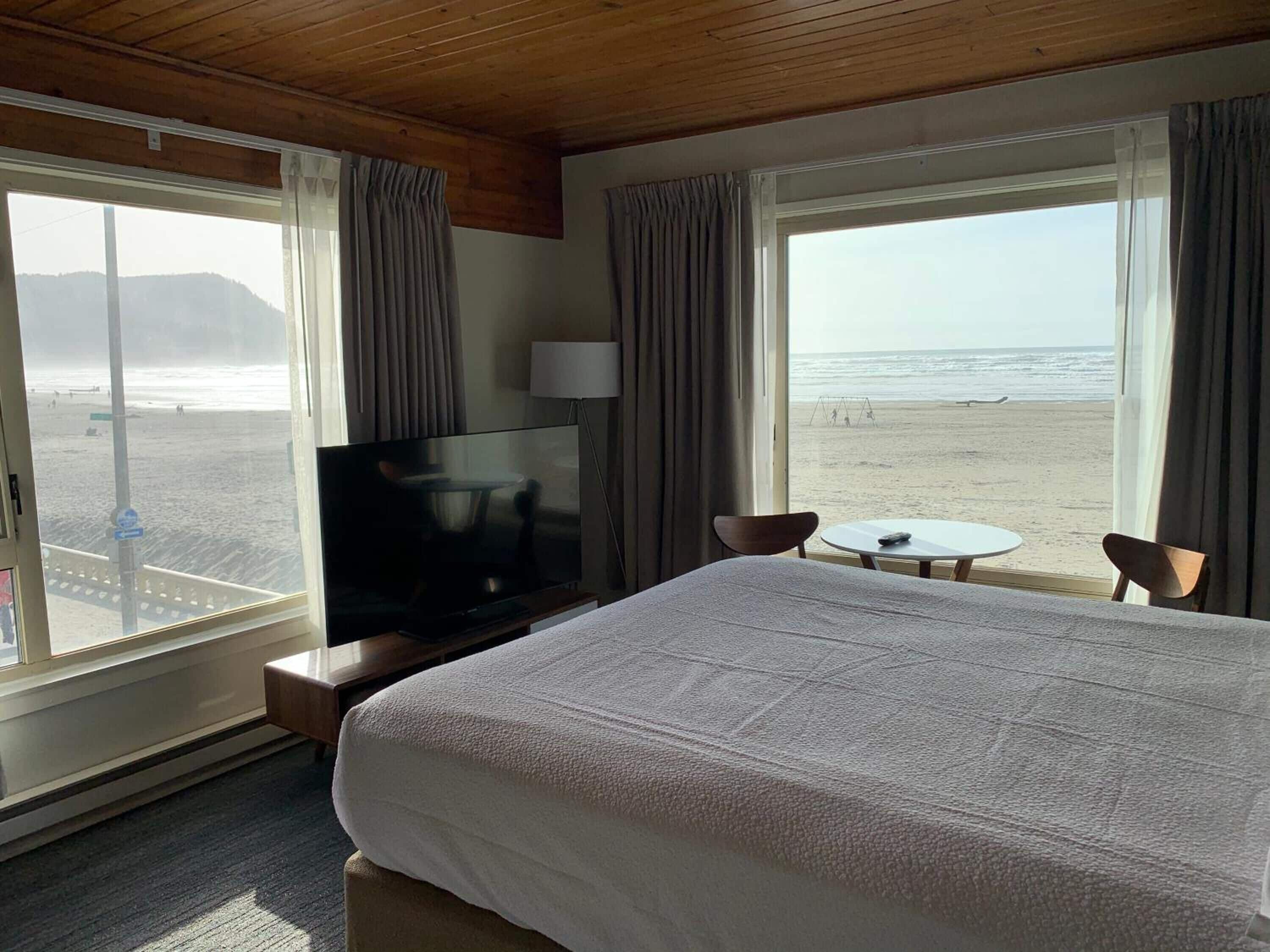 Habitación de hotel con una cama grande, televisor, una mesita y dos sillas. Se ven dos ventanas con vistas a la playa.