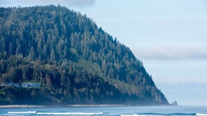 A coastal landscape with a forested hill, a beach, and a lighthouse in the distance. - The Ocean Front at Seaside (Seaside)