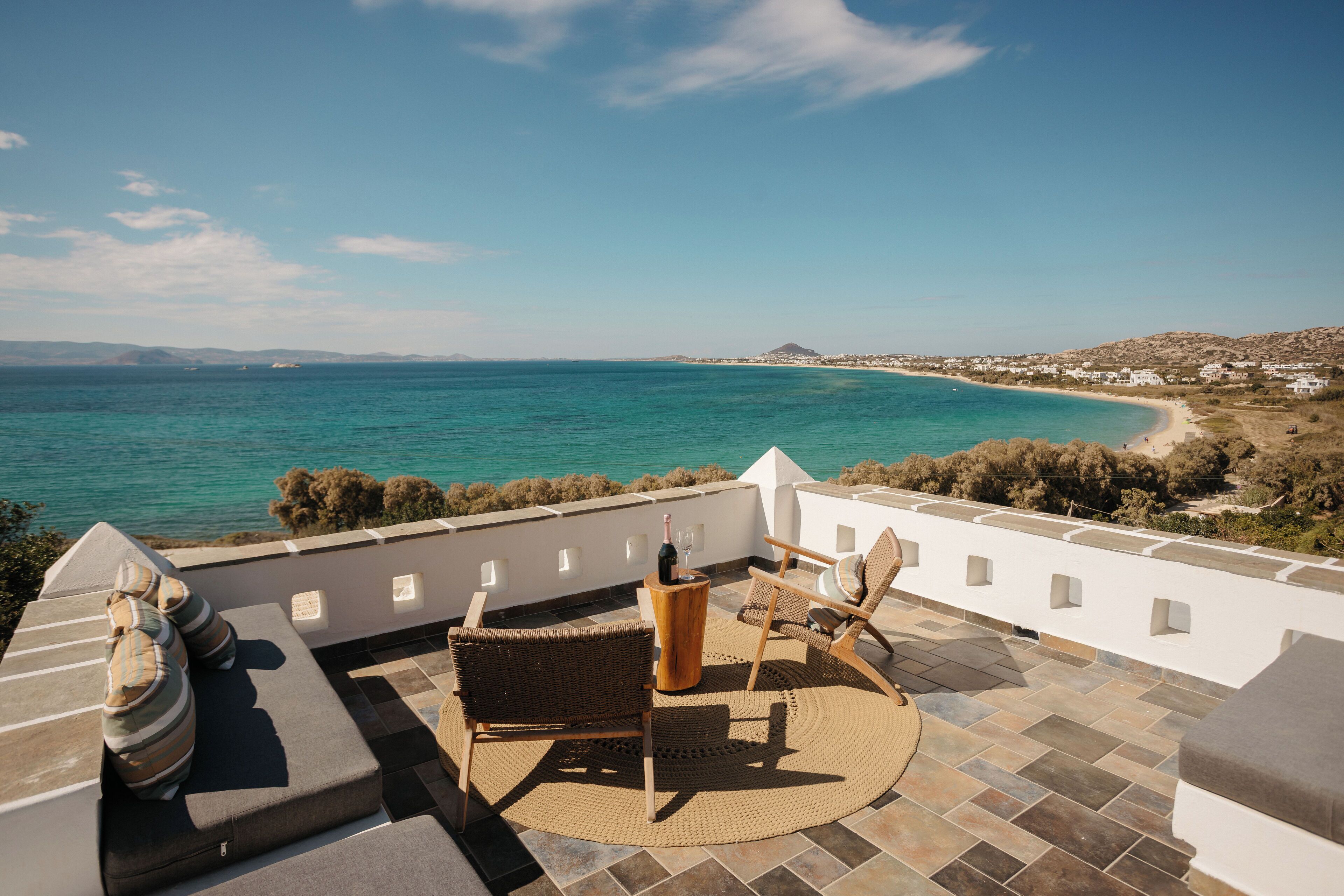 Una terraza con vista al mar, una silla de mimbre, una mesita con una botella y un cojín.
