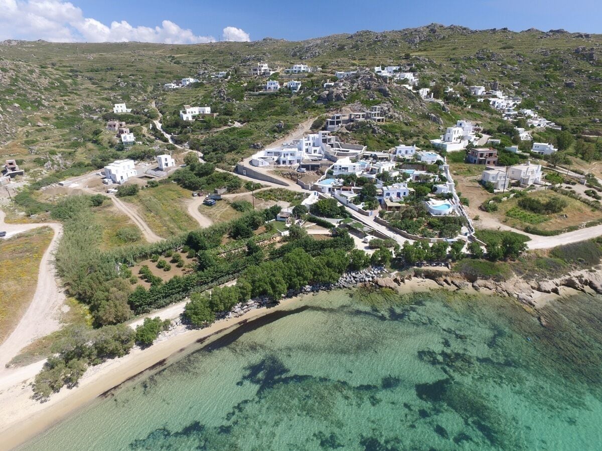 An aerial view of a coastal village with white buildings, clear turquoise waters, and green hills.