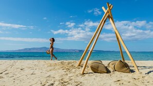 A beach scene with a clear sky, a person walking, and a wooden structure with woven baskets. - Villa Paradise (Naxos)