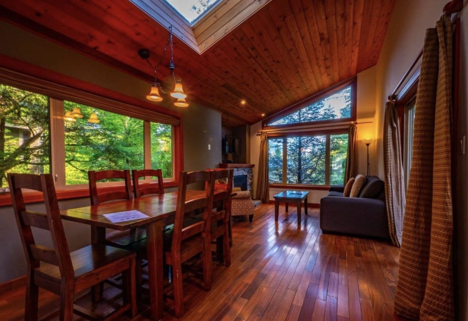 A cozy interior with a wooden dining table, chairs, a sofa, and large windows offering a view of the forest.