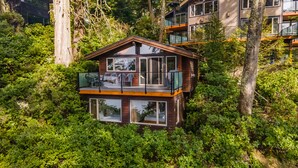 Una casa moderna de dos pisos rodeada de abundante vegetación, con amplias ventanas y un balcón.