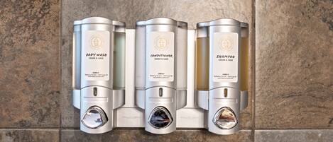 Three cylindrical dispensers on a tiled wall, labeled 'Body Wash', 'Conditioner', and 'Shampoo', each with a similar design and branding.