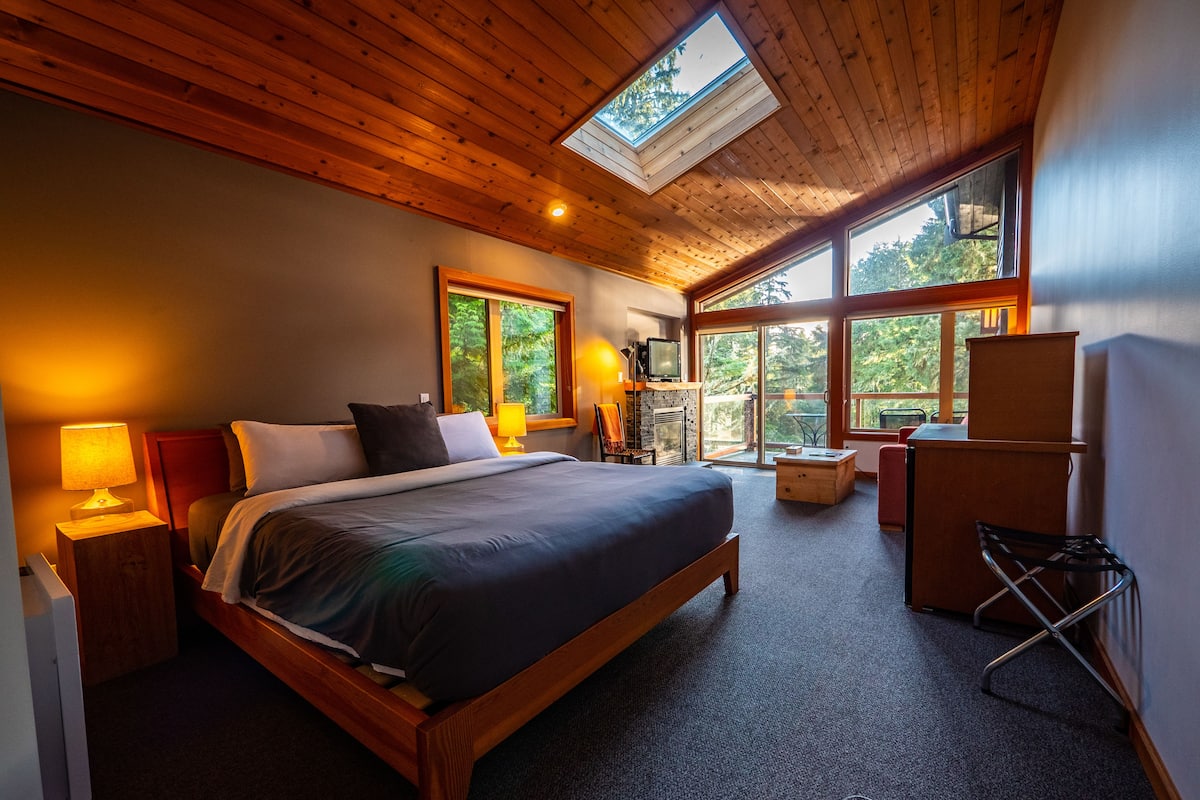 A spacious bedroom with a large bed, a TV, a fireplace, and a balcony with a view of trees.