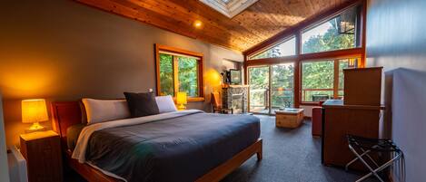 A spacious bedroom with a large bed, a TV, a fireplace, and a balcony with a view of trees.