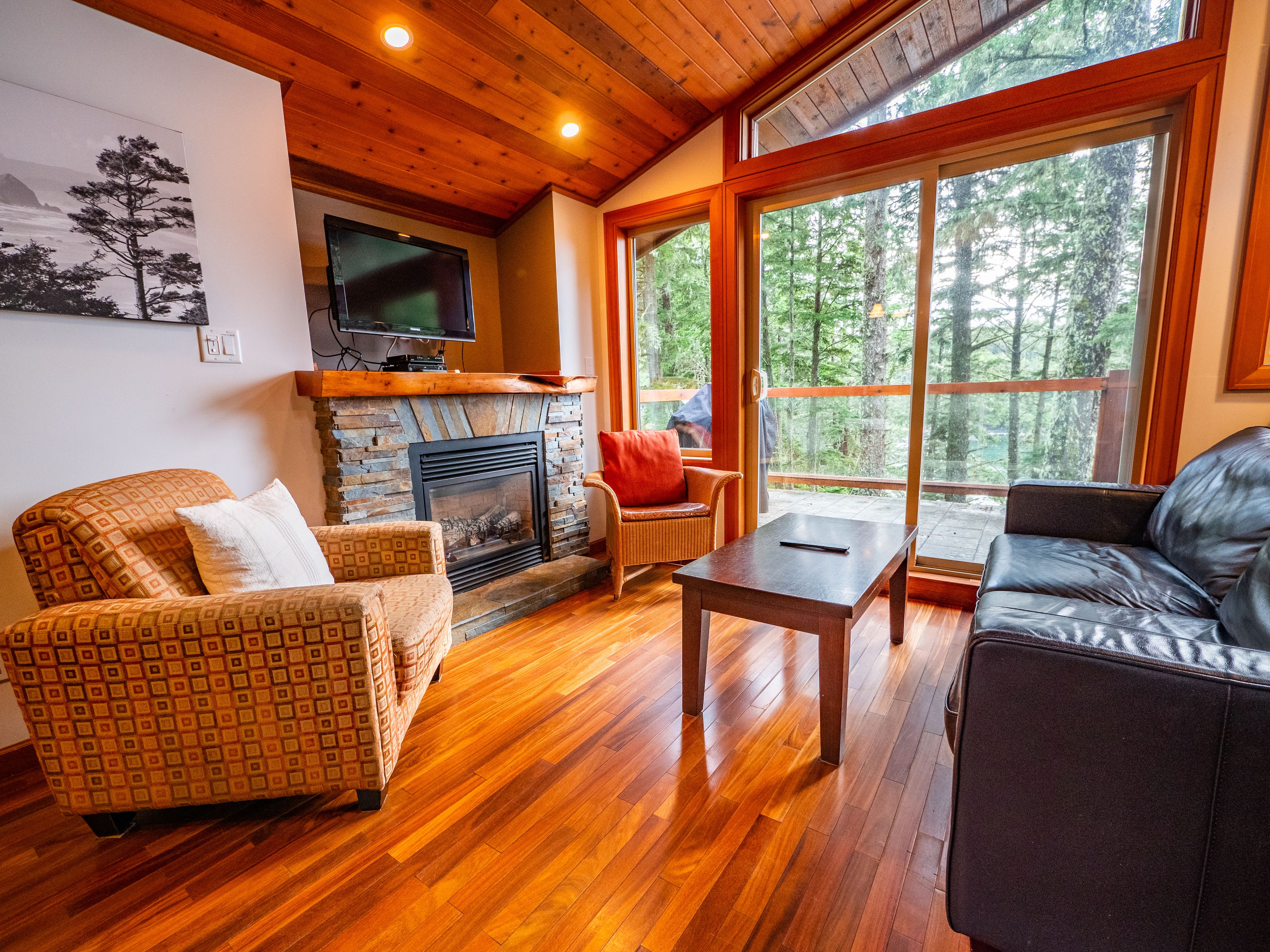 A cozy living room with a fireplace, wooden floors, and large windows offering a forest view.