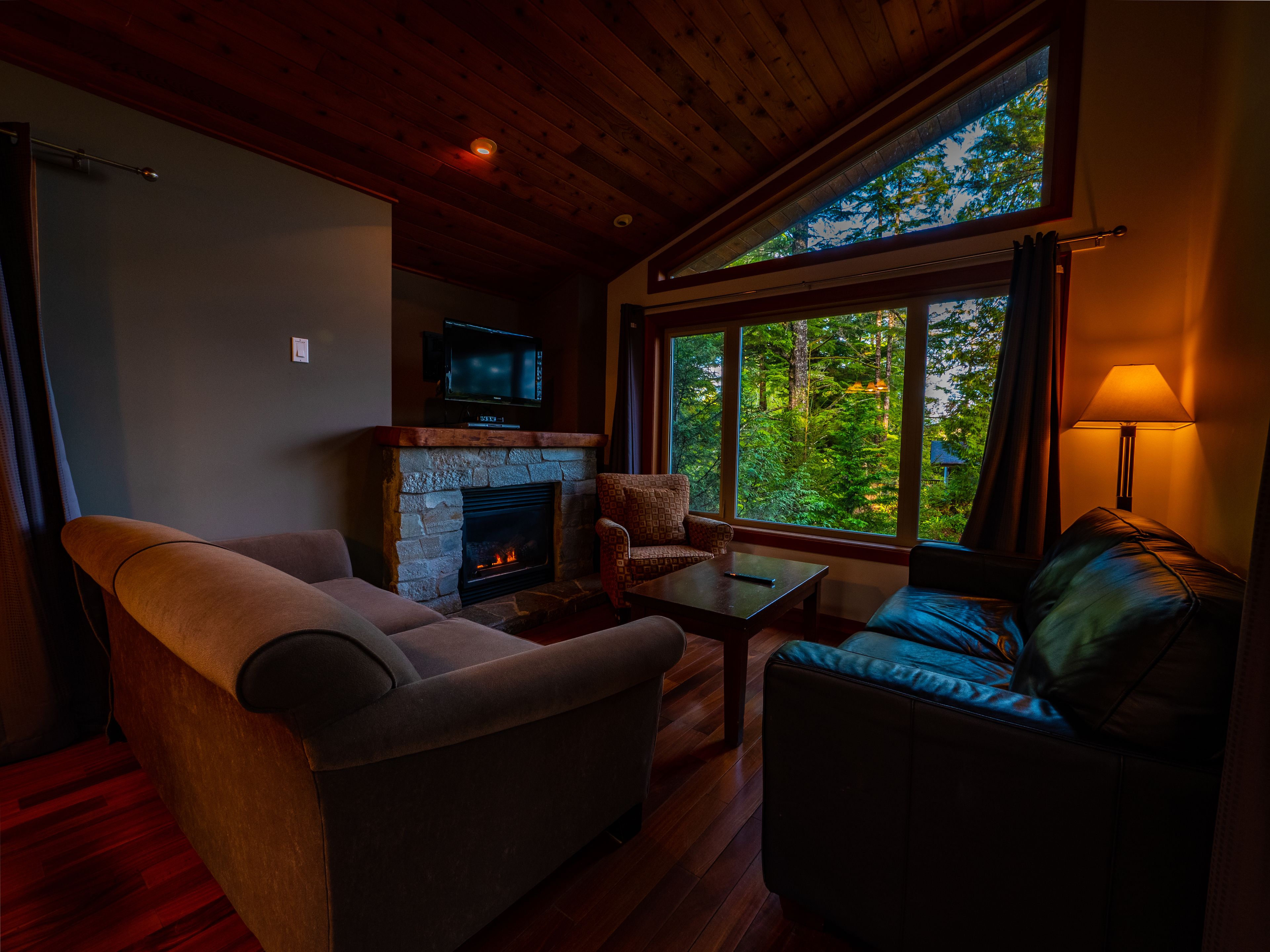 A cozy living room with a stone fireplace, comfortable seating, a television, and a large window offering a view of the forest.
